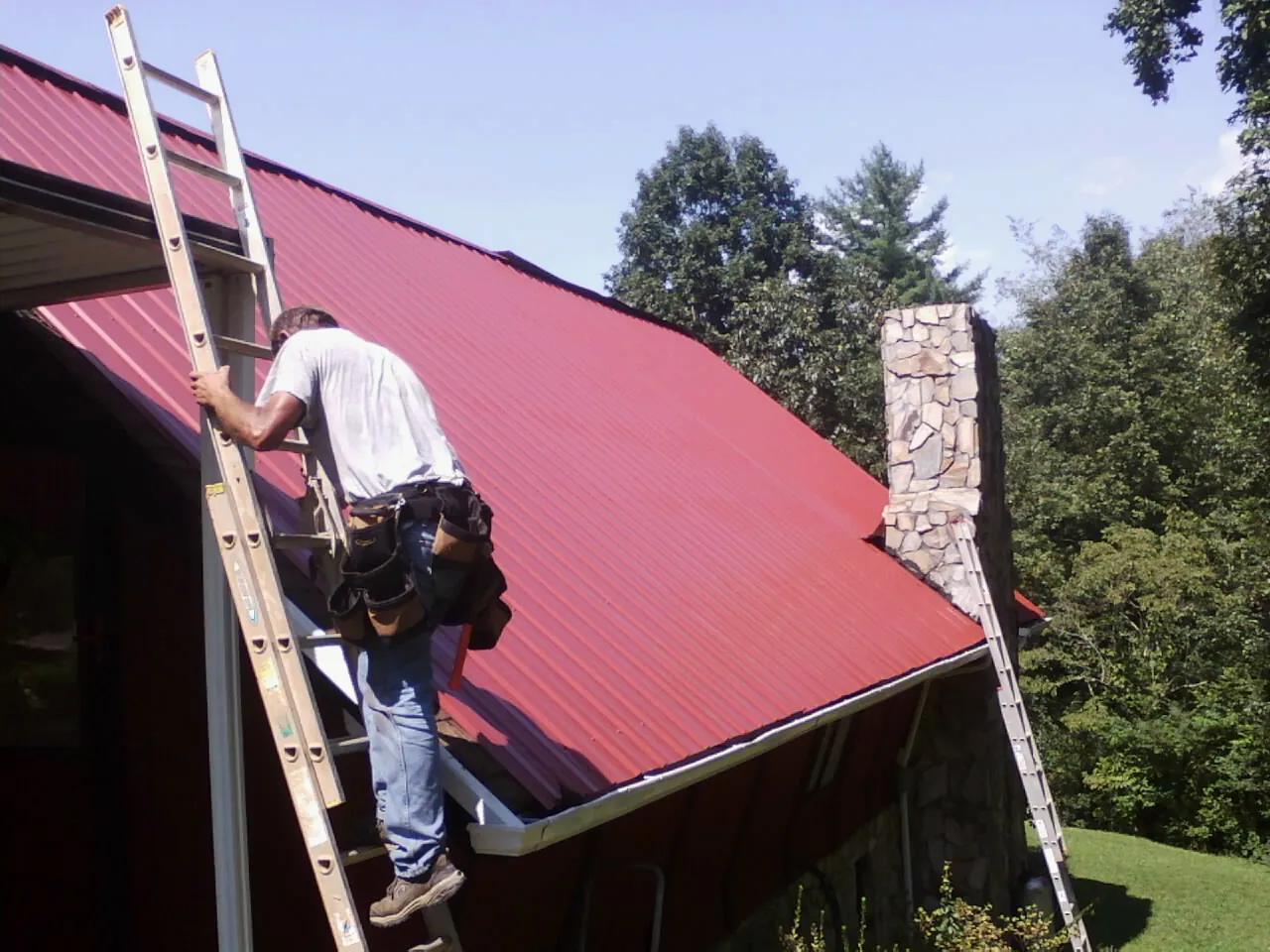 Roofer on a ladder beside a red metal roof and stone chimney — Soffit & Fascia Repair by Roofing Brothers in Playa Vista